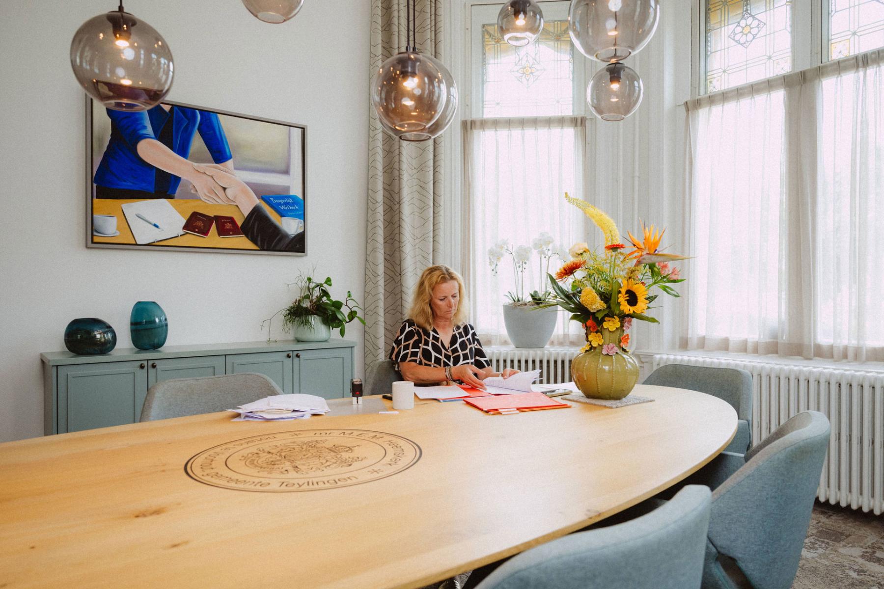 Vrouw zit aan een grote ovale tafel waar een bloempot met zonnebloemen in zitten. De vrouw is bezig met verschillende documenten. Op de achtergrond staat een blauw kastje met drie potten erop en hierboven hangt een schilderij.