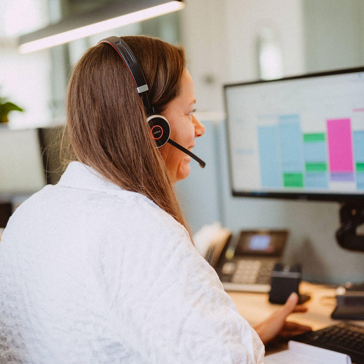 Achter aanzicht van een vrouw met bruin haar die met een headset op zit terwijl ze naar haar scherm kijkt, op haar computerscherm is een agenda te zien.