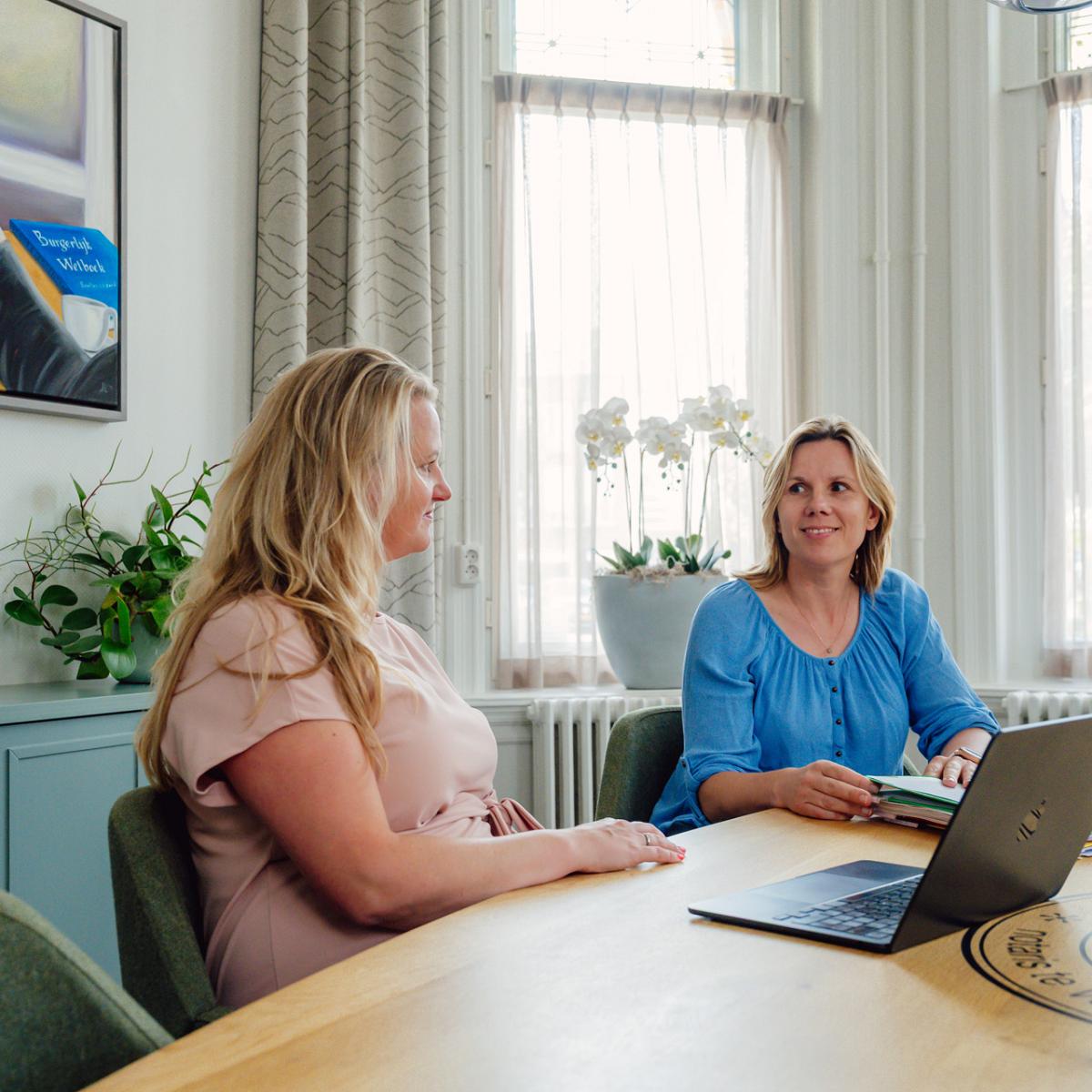 Twee notarismedewerkers in gesprek aan vergadertafel op notariskantoor, met laptop en dossiers in een lichte kantoorruimte