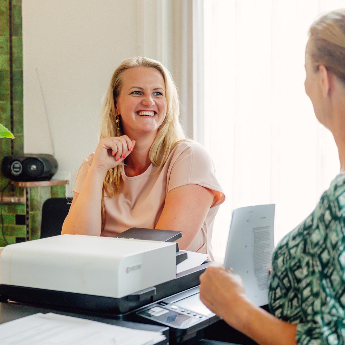 Twee notarismedewerkers in gesprek aan vergadertafel op notariskantoor, met laptop en dossiers in een lichte kantoorruimte