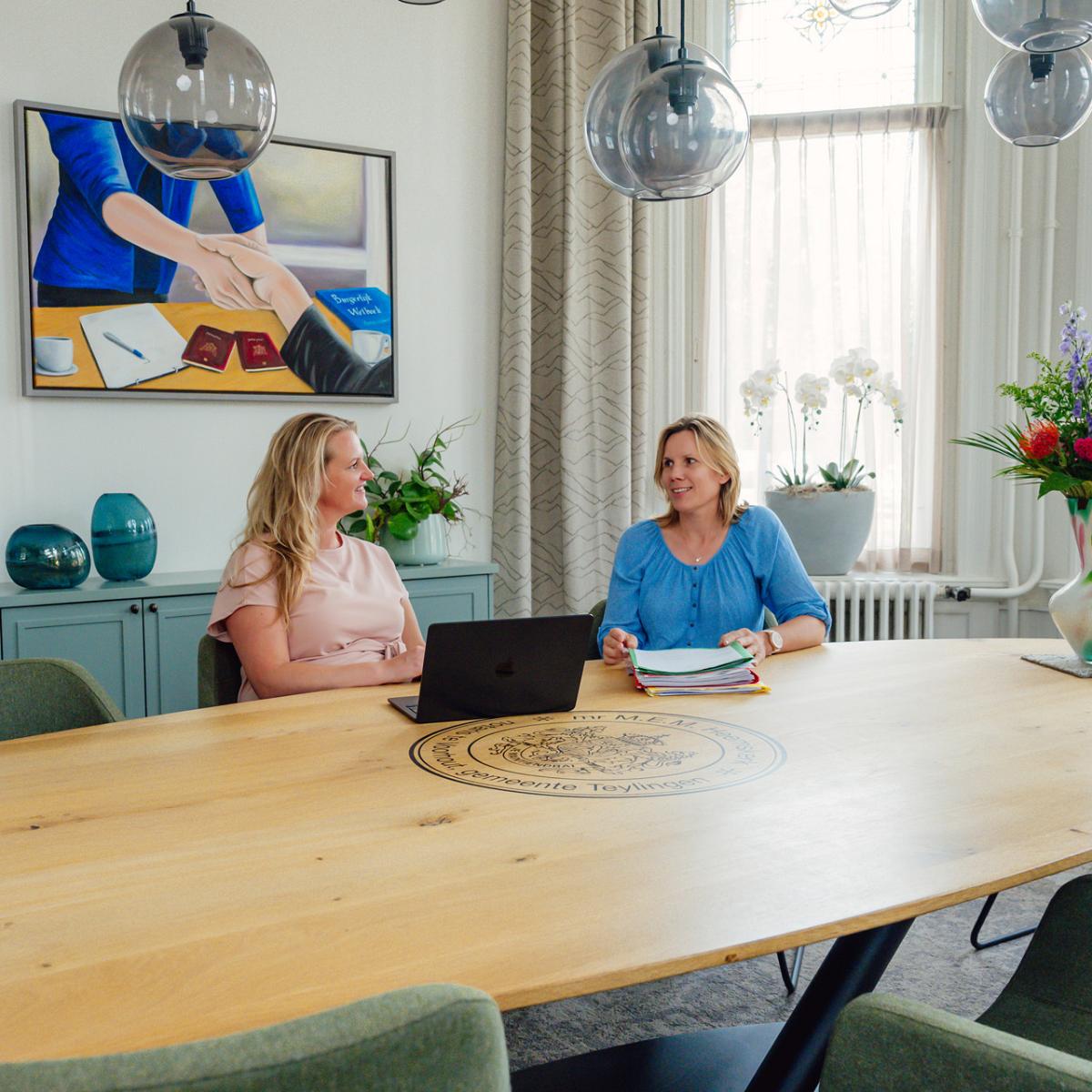 Twee notarismedewerkers in gesprek aan vergadertafel op notariskantoor, met laptop en dossiers in een lichte kantoorruimte