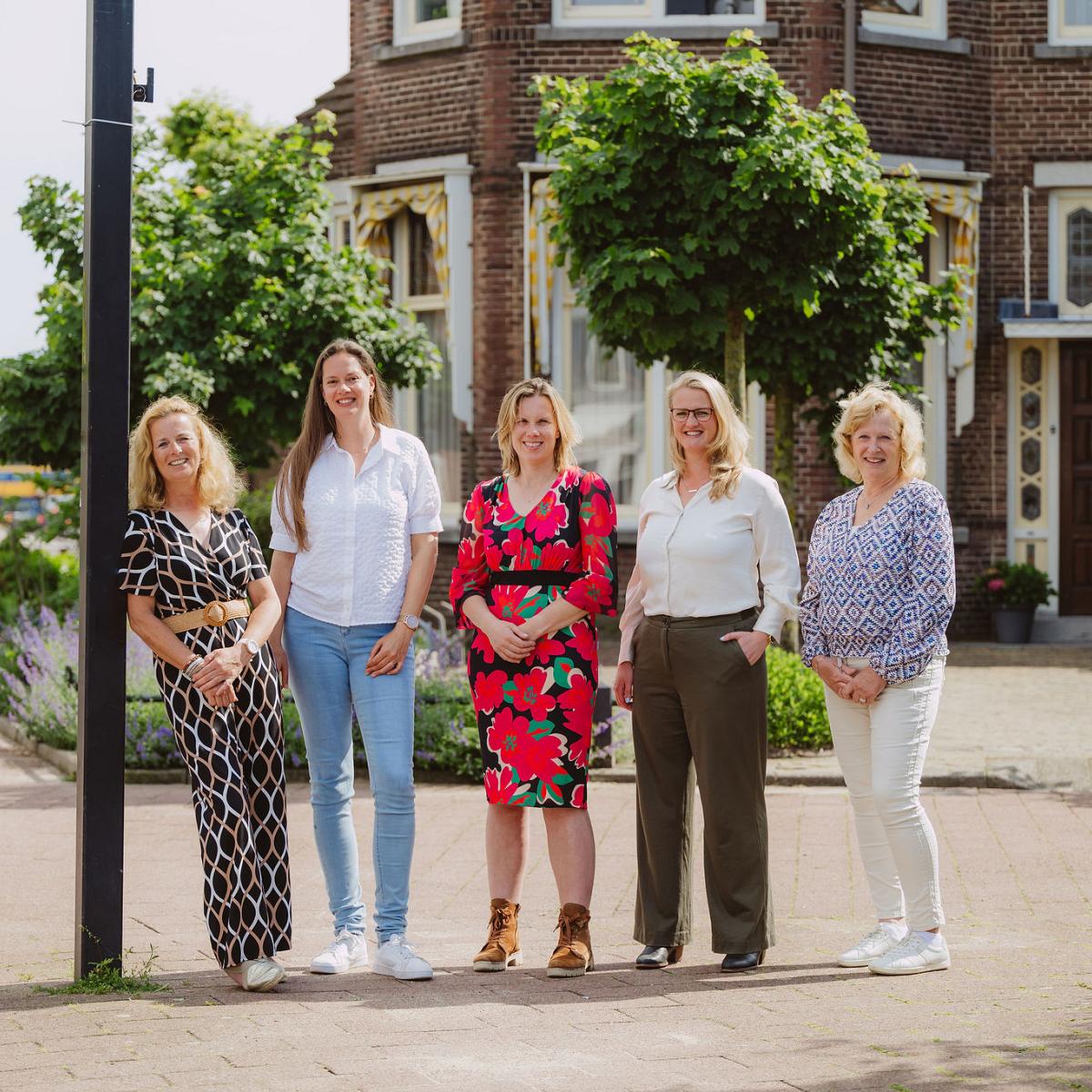 Vijf vrouwen poseren glimlachend voor het bedrijfspand van Notariskantoor Voorhout, omgeven door groene bomen en planten.